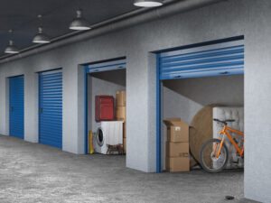 A row of storage units with blue roll-up doors, two of which are open. The open units are filled with items, including a washing machine, red suitcase, cardboard boxes, a bicycle, and a sofa cushion. The area is dimly lit with industrial lighting fixtures above.