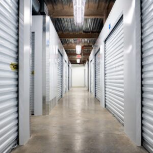 A clean, well-lit hallway in a self-storage facility features rows of silver metal roll-up doors on each side, with a concrete floor and exposed ceiling pipes. An exit sign is visible at the end of the corridor.